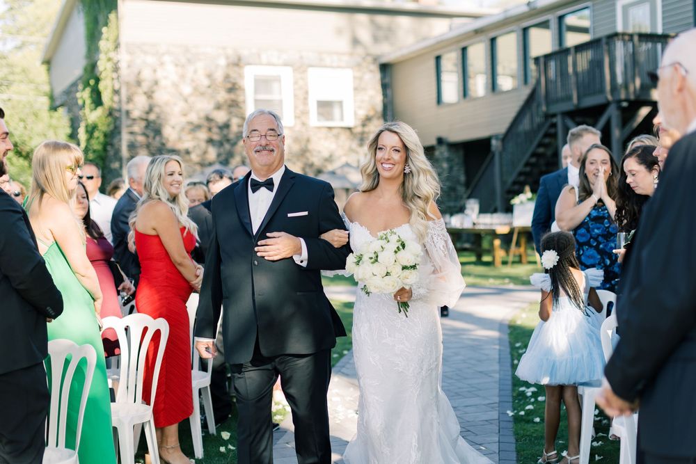 Two people walk down the aisle at an outdoor wedding ceremony while guests look on from white chairs.