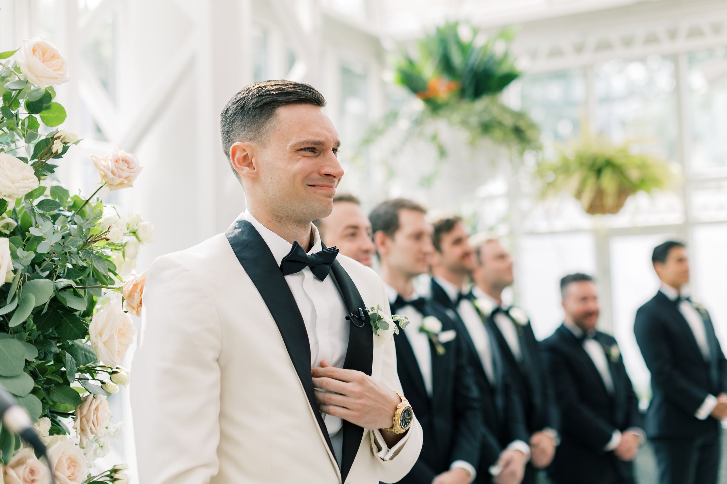 Wedding party stands at altar with groom in white tuxedo next to floral arrangements during elegant ceremony.