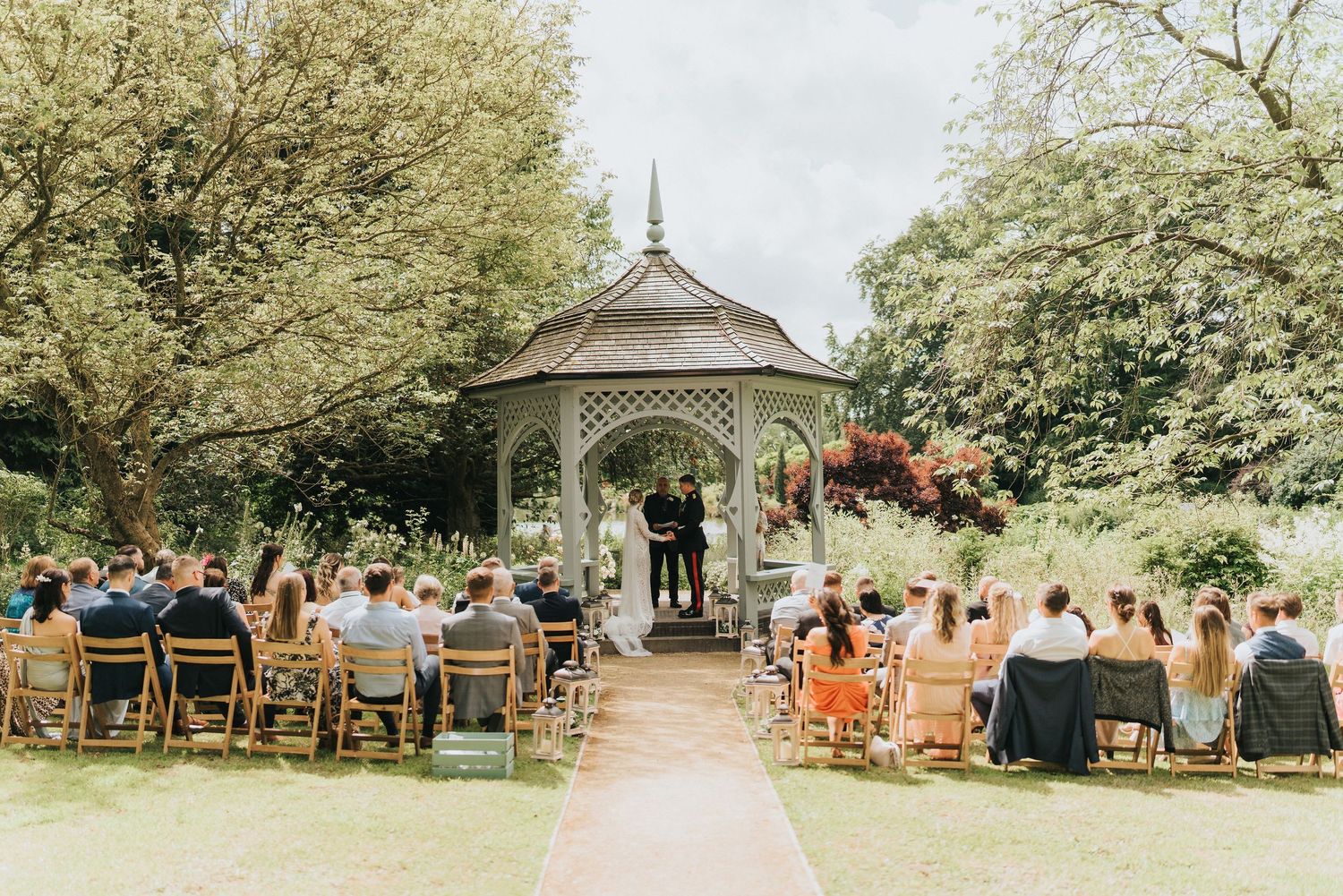 Wedding ceremony takes place at a quaint outdoor gazebo surrounded by trees with guests seated on wooden chairs at Elsham Hall.