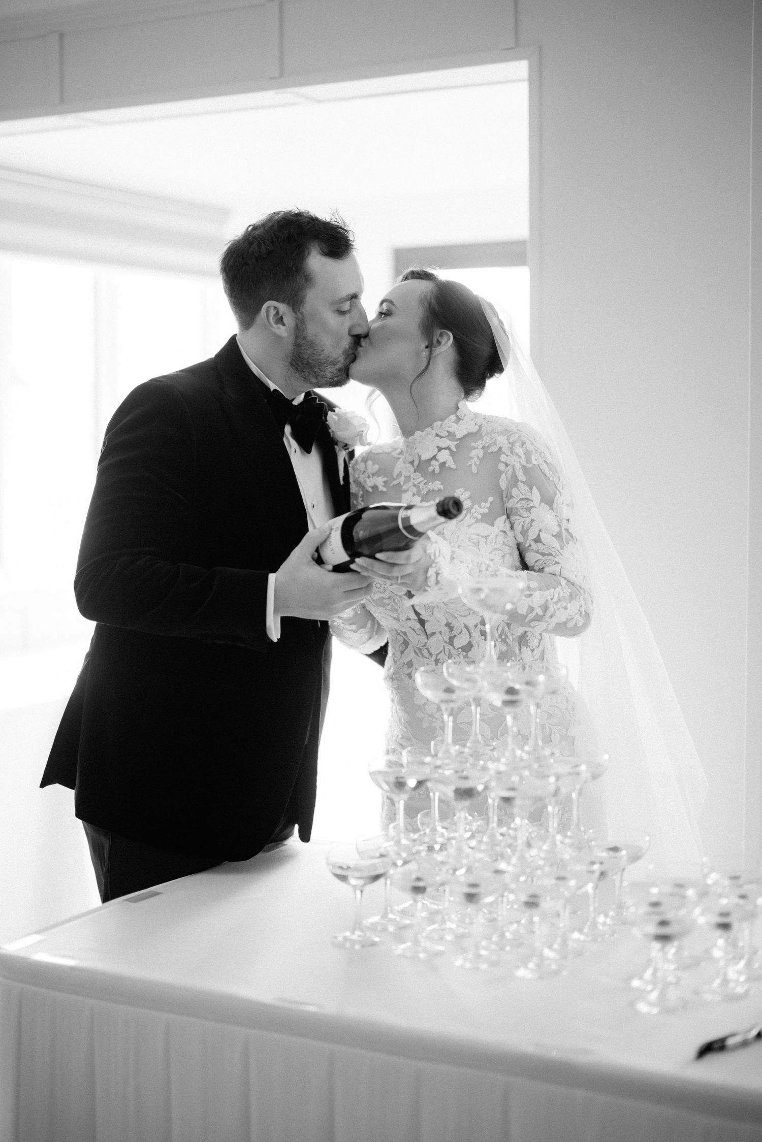 A newlywed couple shares a romantic kiss by a champagne tower at their wedding reception in black and white - taken by Grimsby photographer.