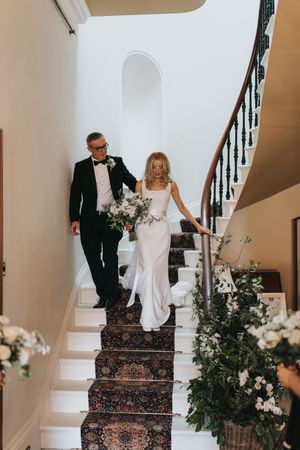A bride and her father descends an elegant staircase decorated with floral arrangements and a patterned carpet runner at Cleatham Hall.