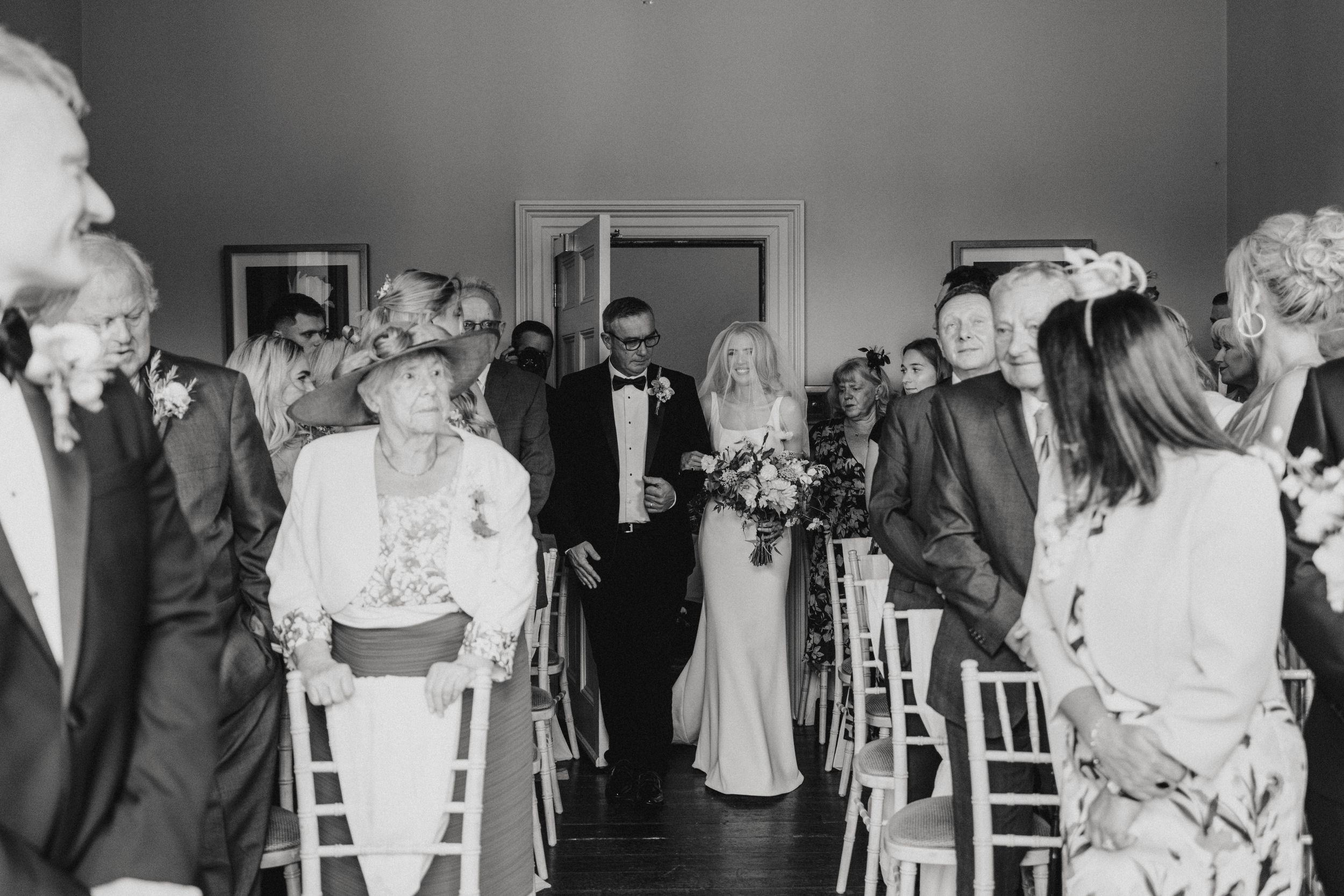 Black and white photo of wedding guests lined up in rows of white chairs watching bride and father walk down the aisle at Cleatham Hall.