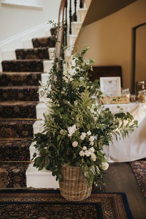 Five identical florals arrangements with white flowers and greenery in wicker baskets line a dark carpeted staircase at Cleatham Hall.