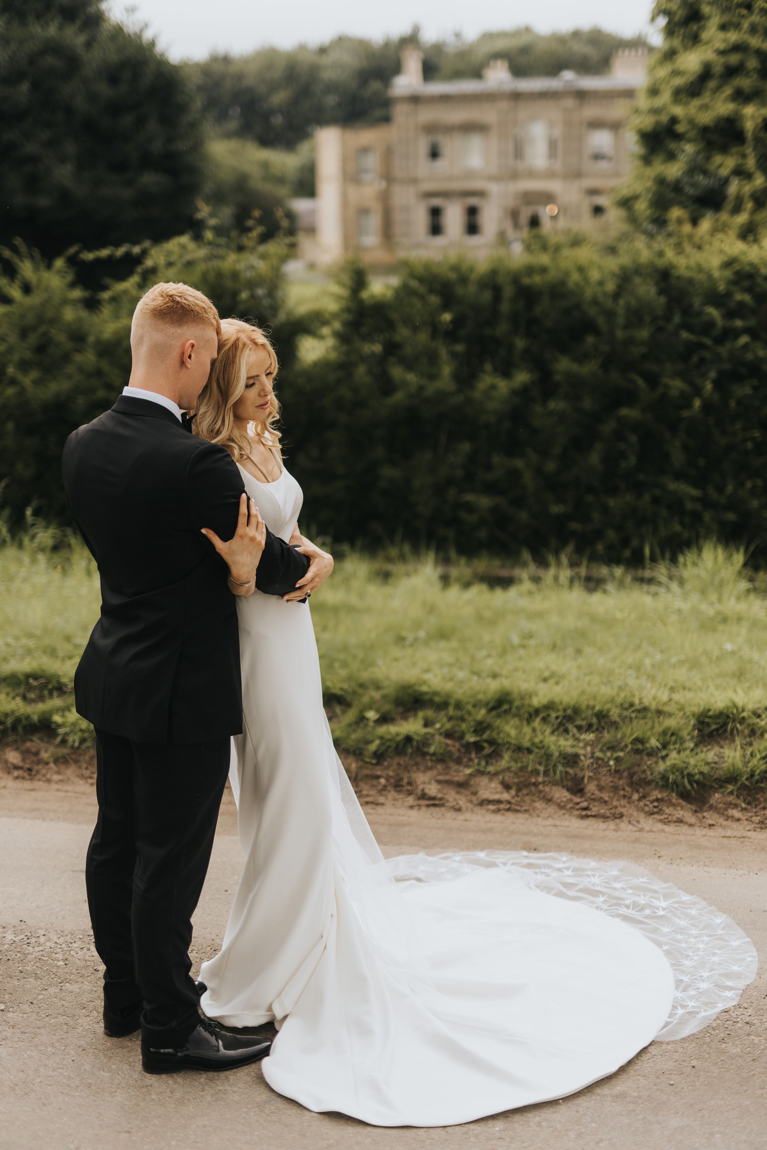 Couple in formal wedding attire embrace on path with Cleatham Hall visible in background.