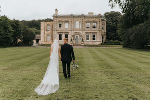 Wedding couple walks across Cleatham Hall grounds with elegant building visible in background.