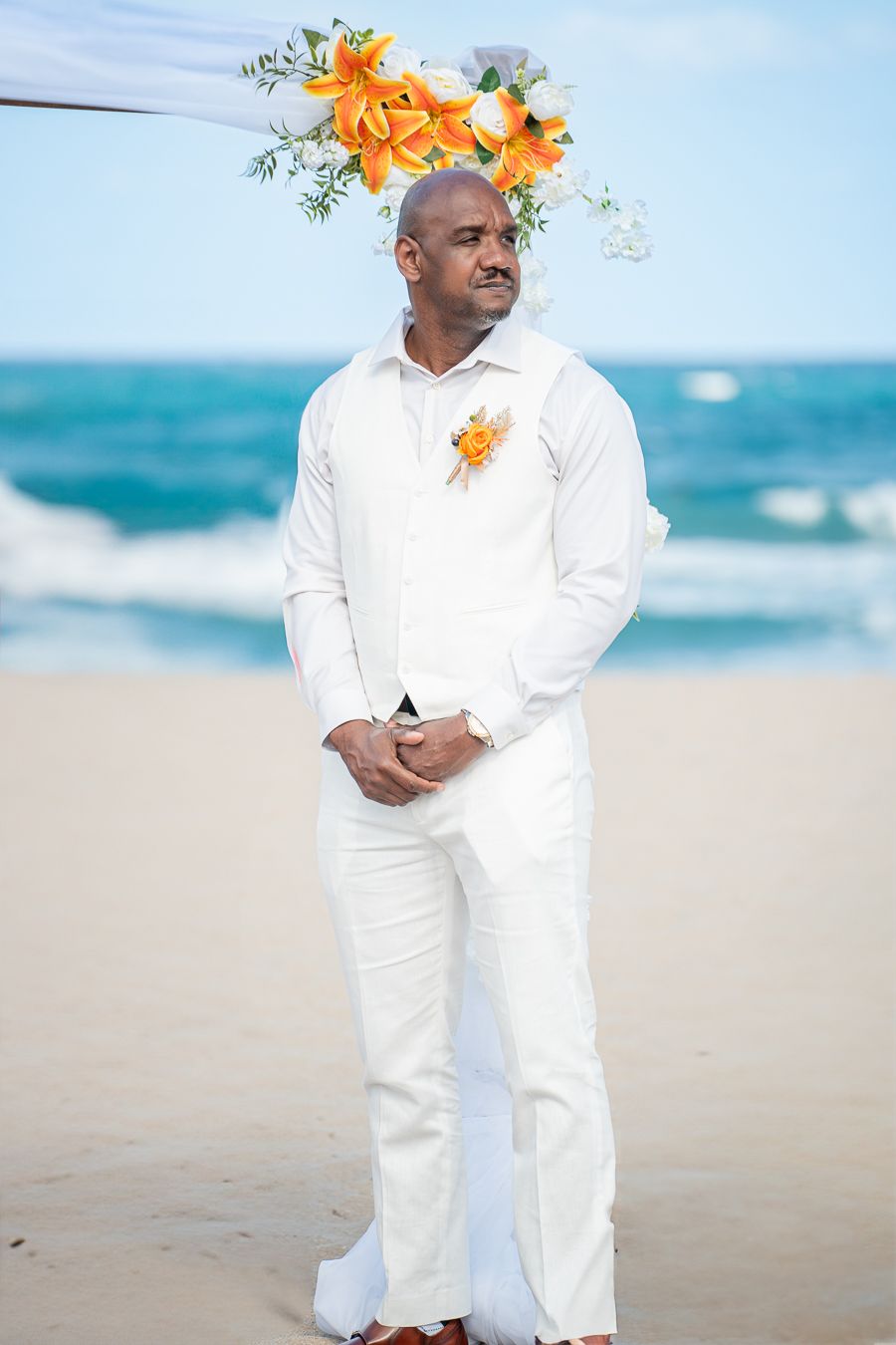 Elegant beach wedding attire featuring an all-white suit with orange floral accents against turquoise ocean backdrop.