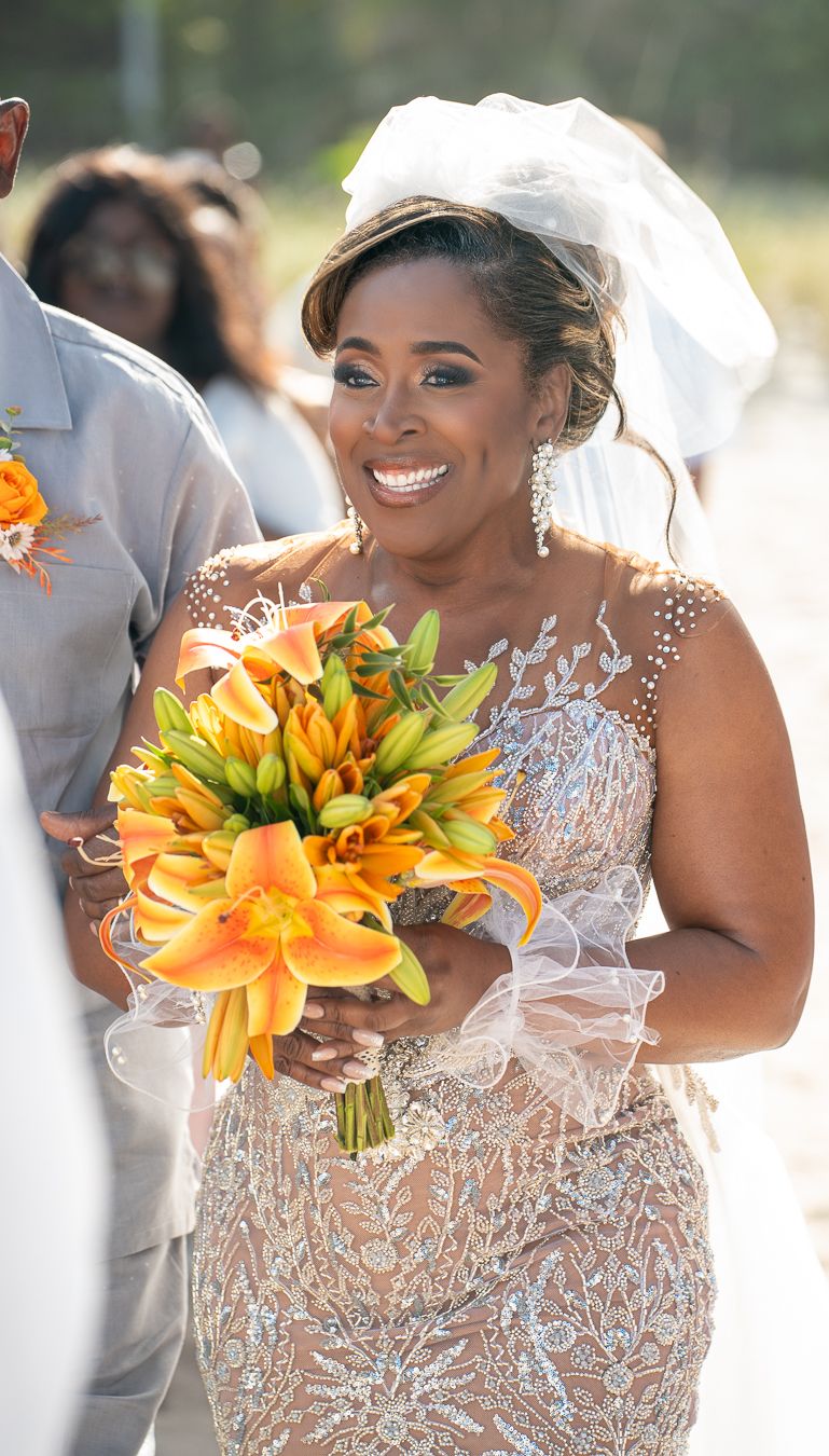 Wedding party member in an elegant beaded dress holds vibrant orange lilies during an outdoor celebration.