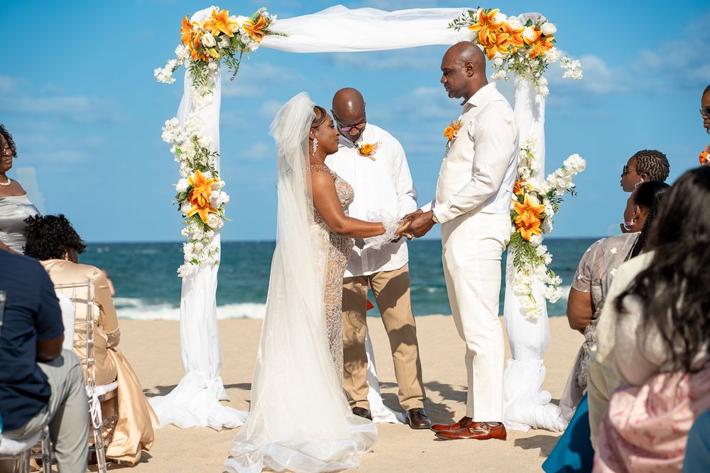A series of people gathered under white floral wedding arches on a beautiful sunny beach during a ceremony.