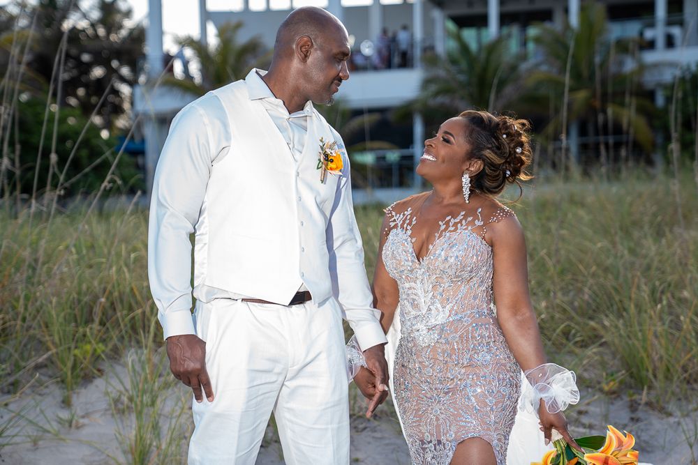 A couple shares a joyful moment together on a sandy beach wearing white and beige formal attire.