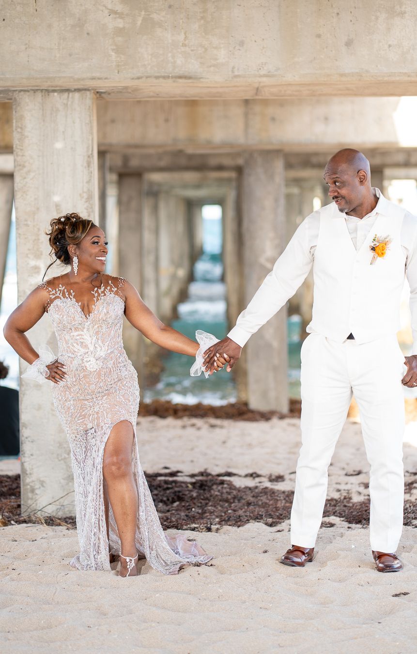 Bride in sparkly white gown holds hands with groom in white suit during romantic beach wedding photo session.