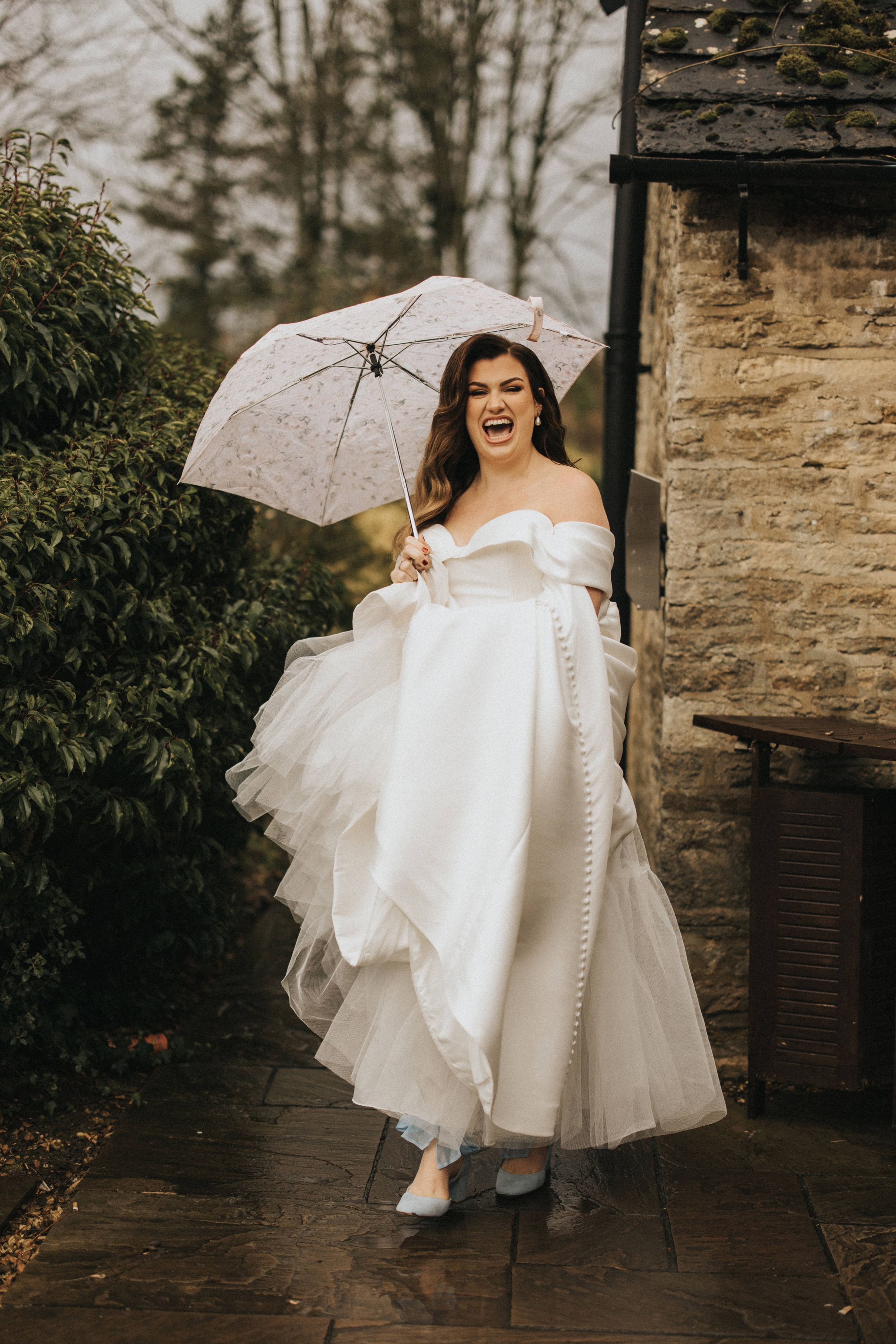 A joyful person in a flowing white wedding dress holds a white umbrella while standing next to a stone building.