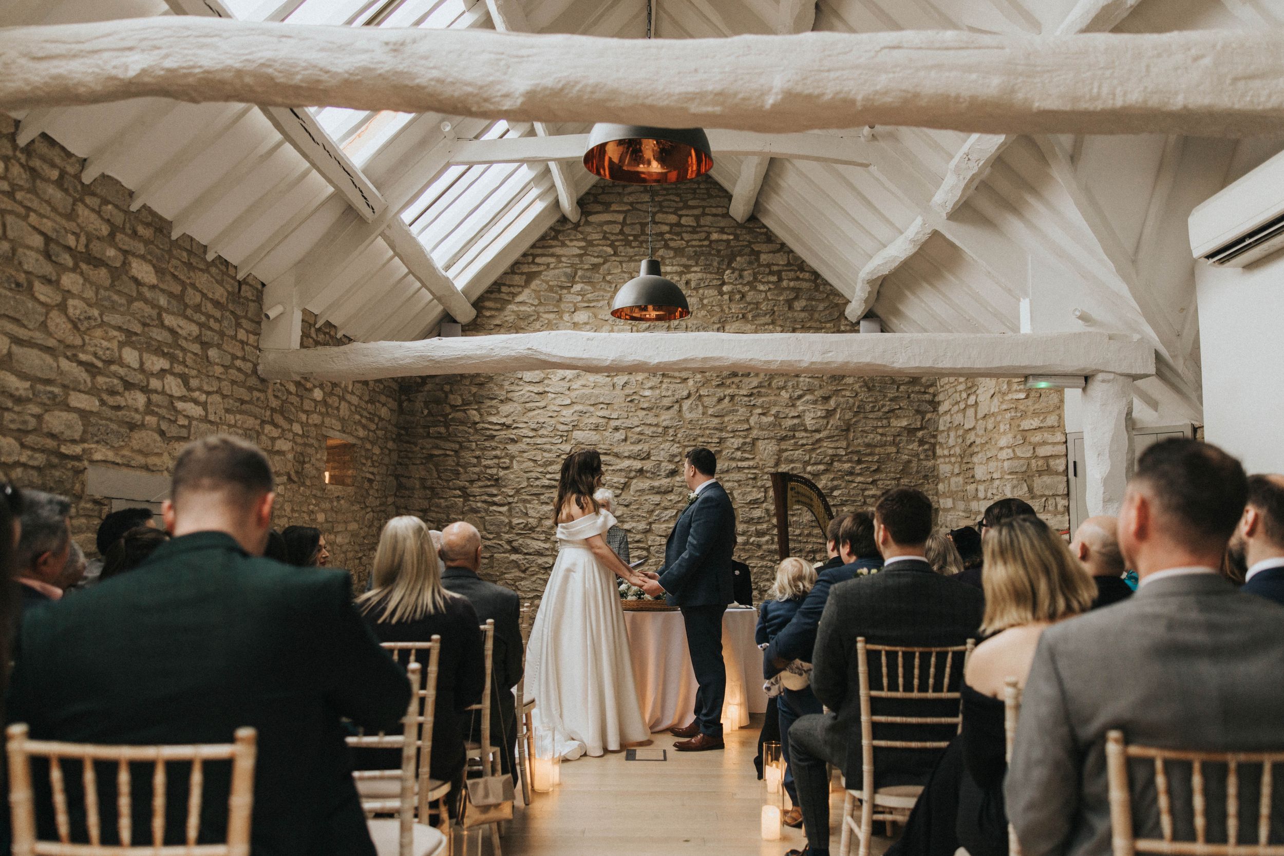 Intimate wedding ceremony taking place in a rustic barn venue with exposed stone walls and white wooden beams.
