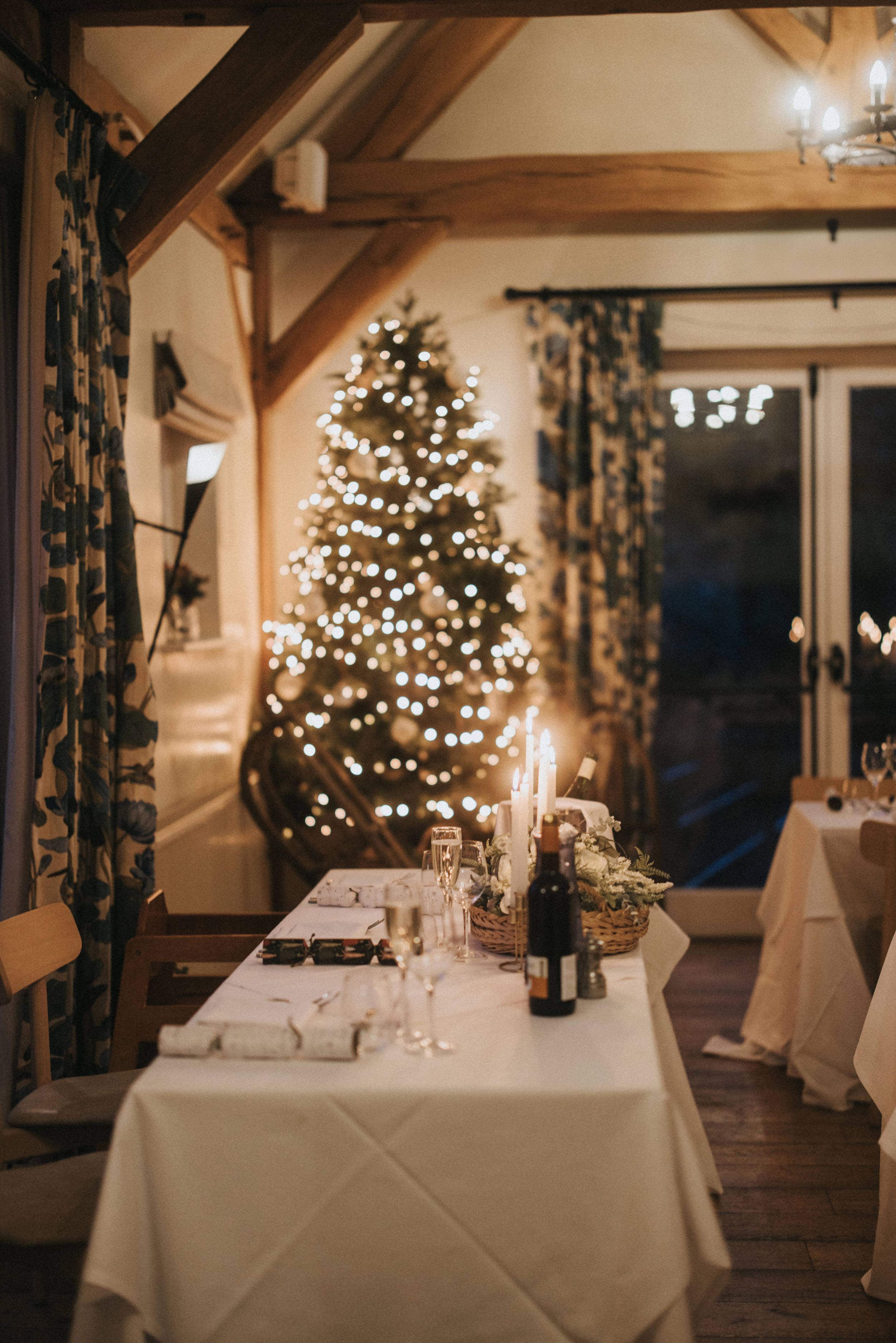 A cozy holiday table setting with candlelight and white linens against a glowing Christmas tree in a rustic room.