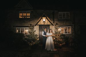 Warmly lit cottage house exterior with glowing windows and decorated Christmas trees at night.