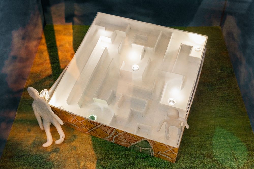 Architectural scale model showing interior layout with miniature figure standing beside illuminated white structure on grass base.