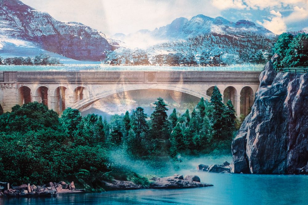 A scenic panorama of a stone viaduct bridge spanning across a tranquil lake surrounded by lush green forest and cliffs.