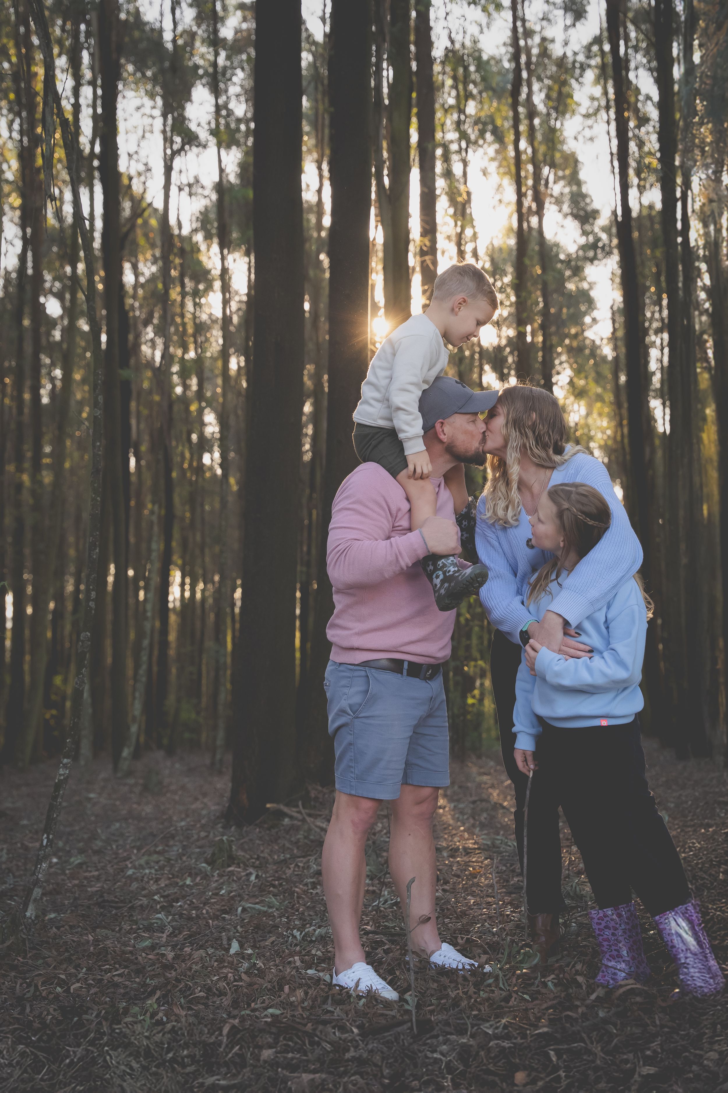 Famlily in a gum forest with dad and son on his shoulder bending to kiss mom and daughter in a family embrace.