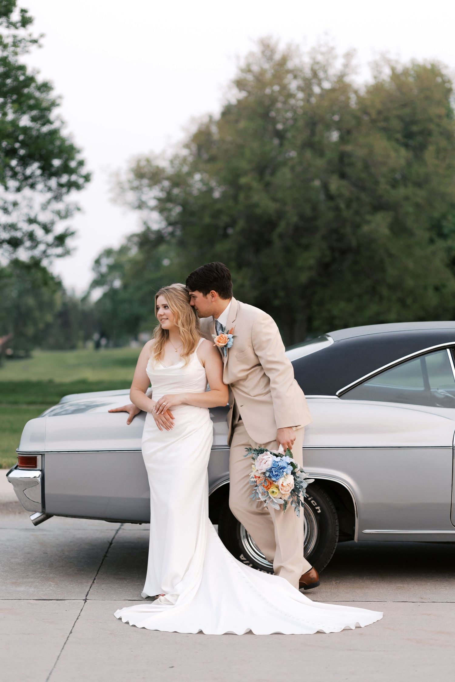 A newlywed couple stands in front of a vintage car and church on their wedding day, wearing tan and white attire.