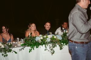 Wedding party seated at a decorated head table with greenery garland decor.