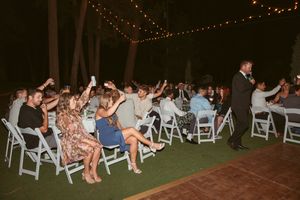 Wedding guests seated in white chairs raise their arms during an outdoor evening reception.