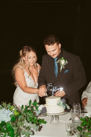 Newlyweds cut their white wedding cake together at a beautifully decorated reception table with greenery.