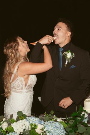 Bride and groom playfully feeding each other cake at their wedding reception with floral decorations.