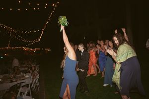 Wedding guest in blue dress catches bouquet toss at outdoor evening reception with string lights.