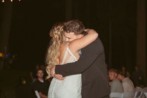 A couple shares their first dance at a dimly lit wedding reception with guests watching in the background.