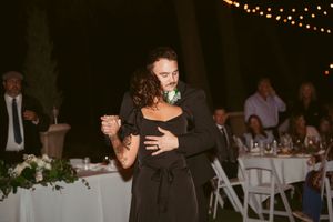A couple slow dances together at an evening wedding reception with string lights and white tables in the background.