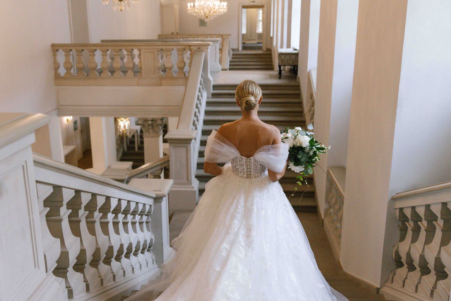 An ornate staircase in a grand interior features elegant white balustrades and a crystal chandelier overhead.
