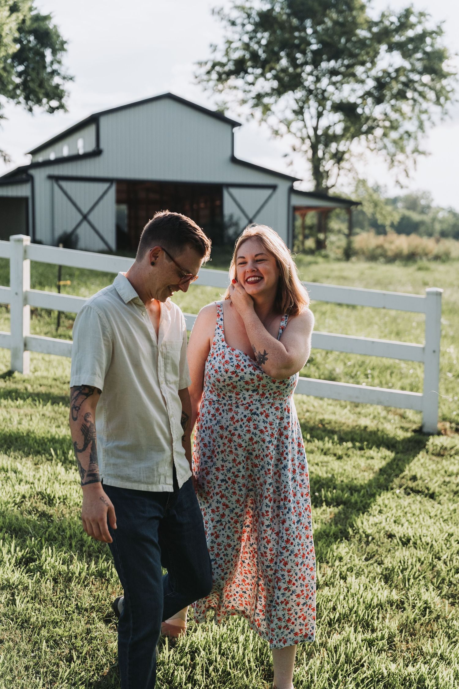 A couple shares a candid moment together while standing on a sunny lawn with a barn and white fence in the background.