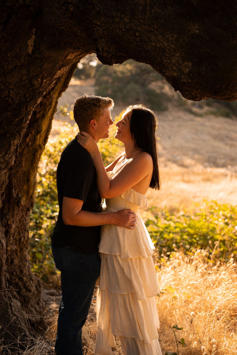 Sweet couple sharing tender moments under oak tree branches during sunset photoshoot.