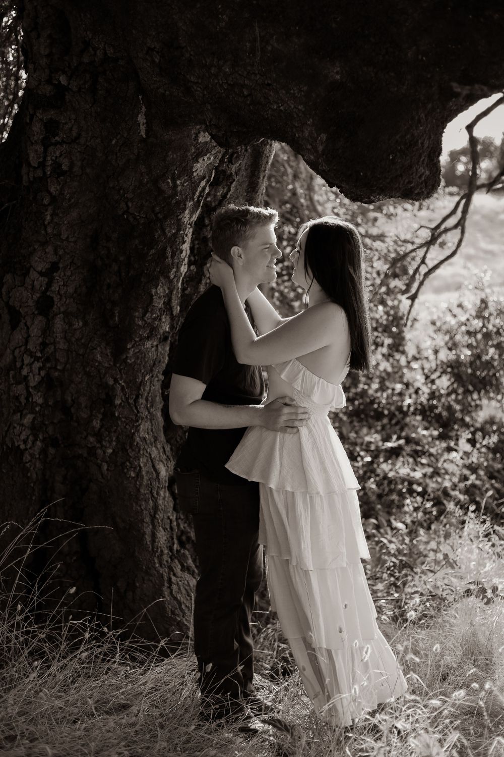 A romantic couple shares a tender embrace against a tree in a dreamy black and white photograph.