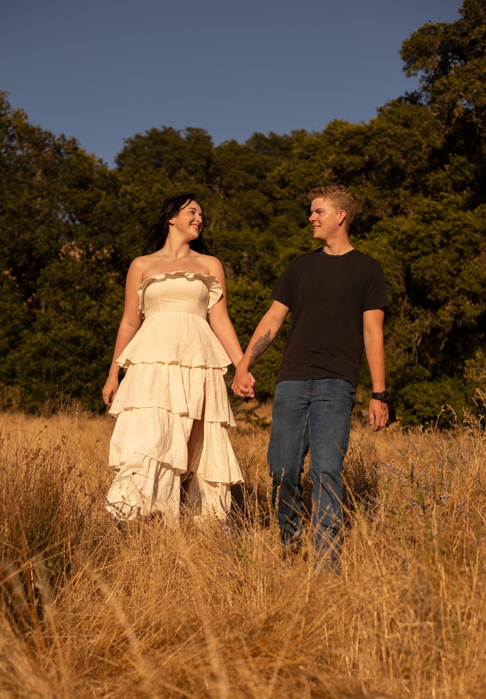 A couple laughs together while holding hands in a sun-drenched field with oak trees in the background.