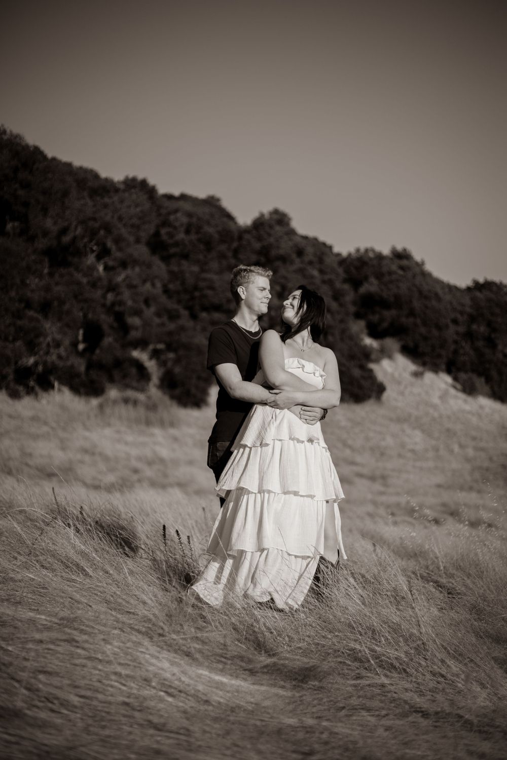 A romantic couple stands together in tall grass with a dreamy sepia-toned effect.
