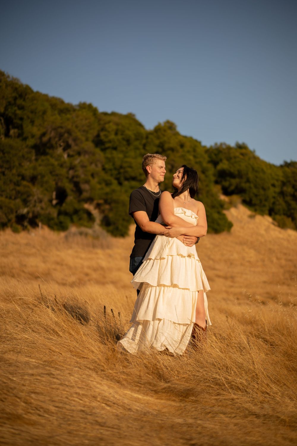 Couple standing together in tall golden grass with hills in background during sunset.