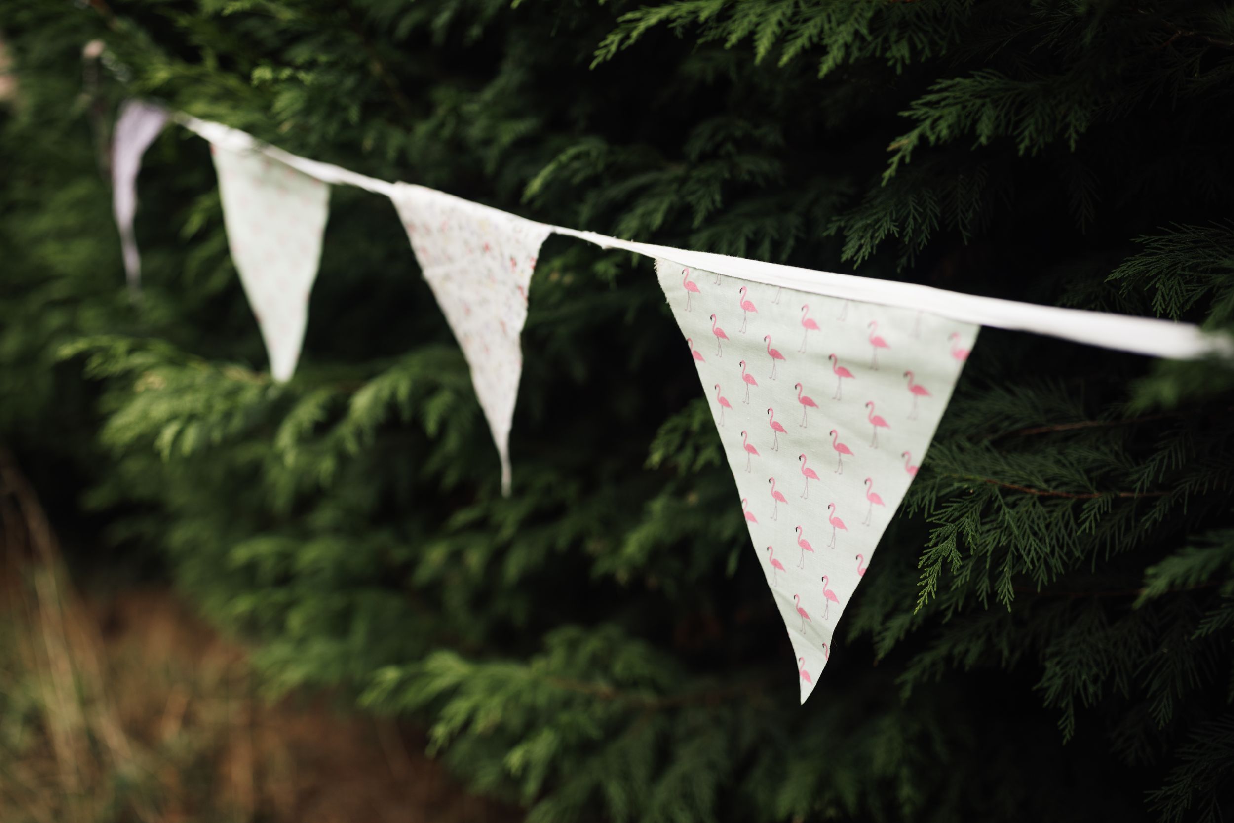 White triangle bunting flags with pink floral patterns hang from string against dark green foliage background.