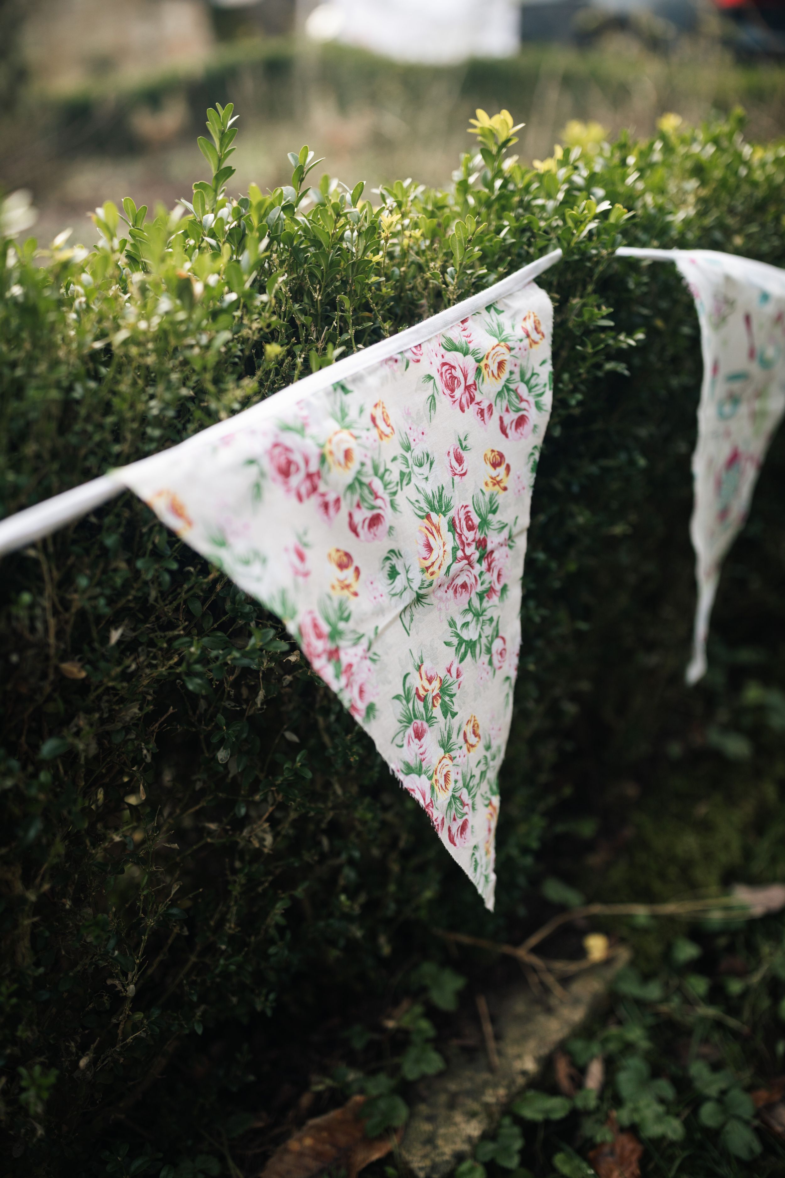 Floral printed fabric bunting with pink and green patterns hangs against a green hedge backdrop.