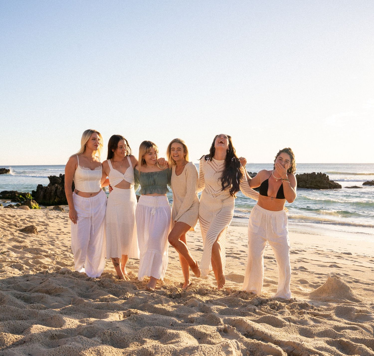 Group of friends in white outfits posing together on a beach at sunset.