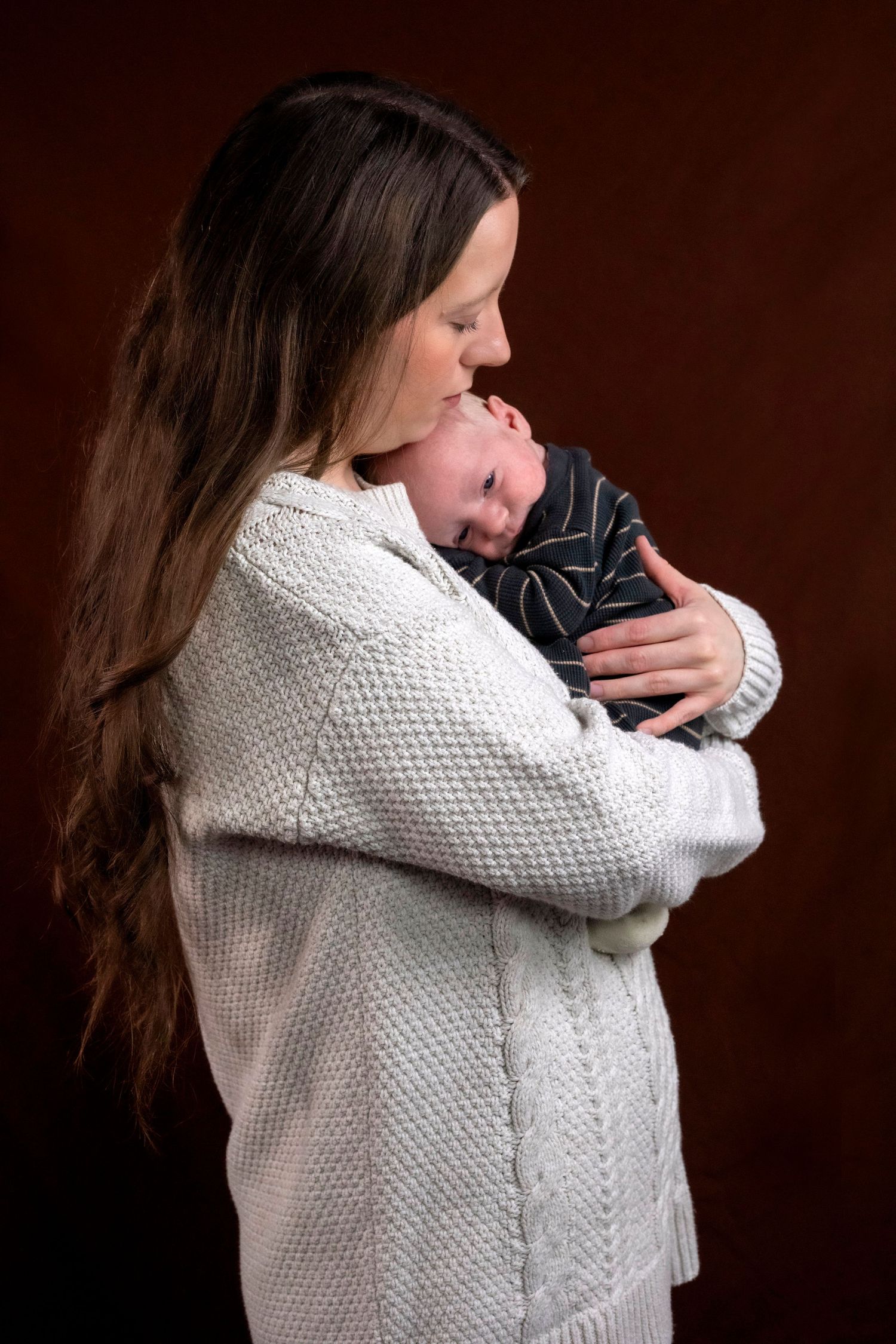 A tender moment as a mother in a white knit sweater cradles and kisses her sleeping newborn baby.