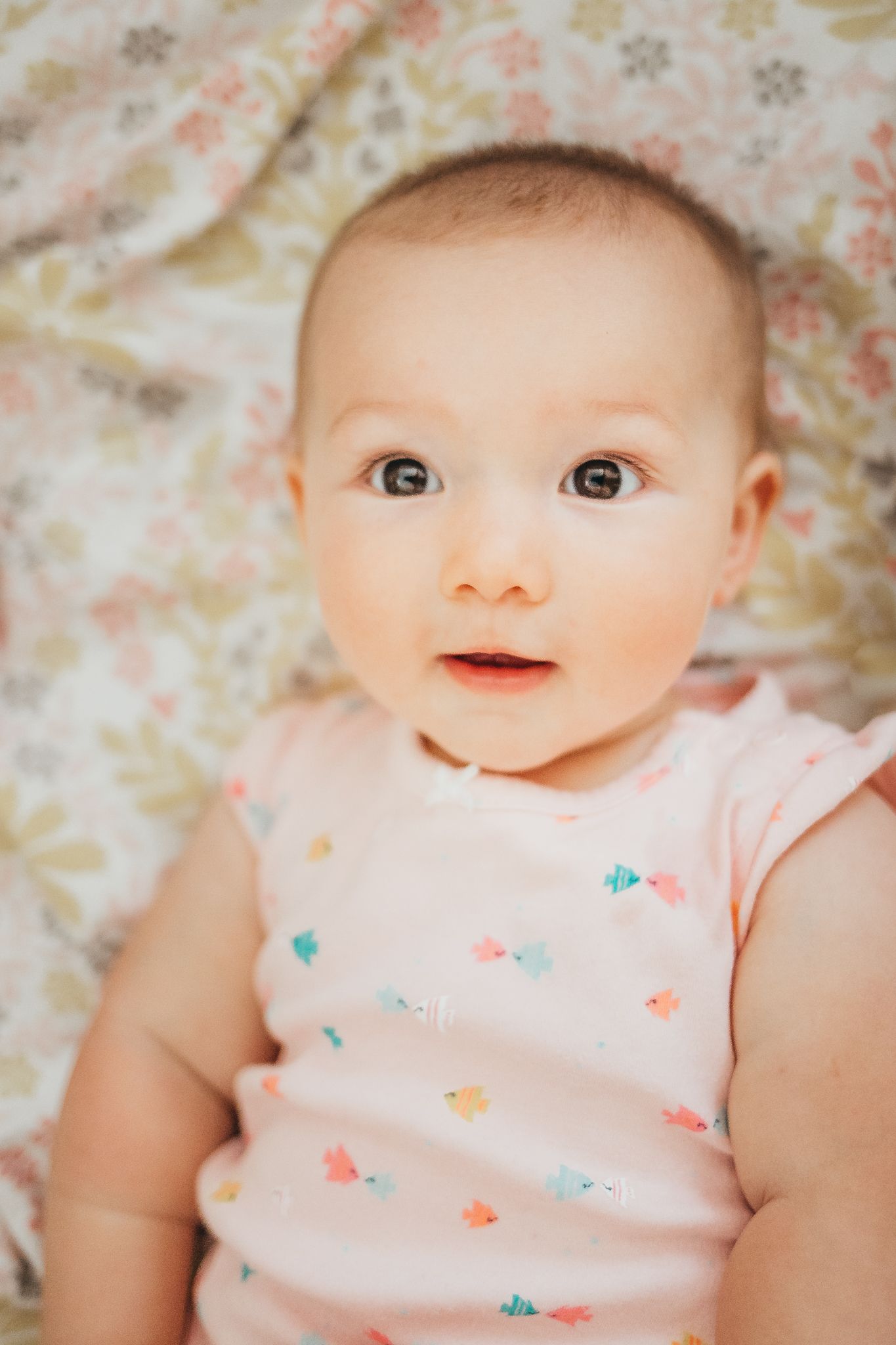 Adorable baby girl in pink floral onesie lying on a floral blanket, looking up with wide curious eyes.
