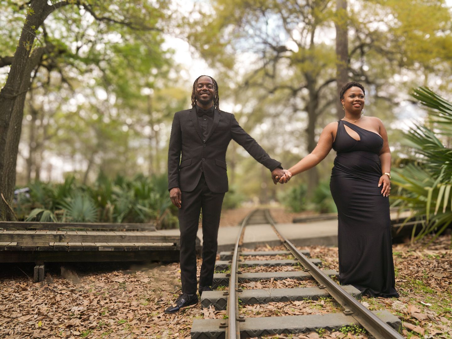 Elegant couple holding hands on railroad tracks in a forest, wearing formal black attire.