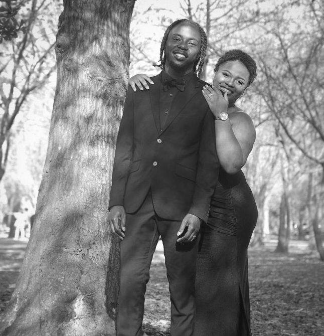 Black and white photo of a smiling couple posing together by a tree in a park, dressed in formal attire.