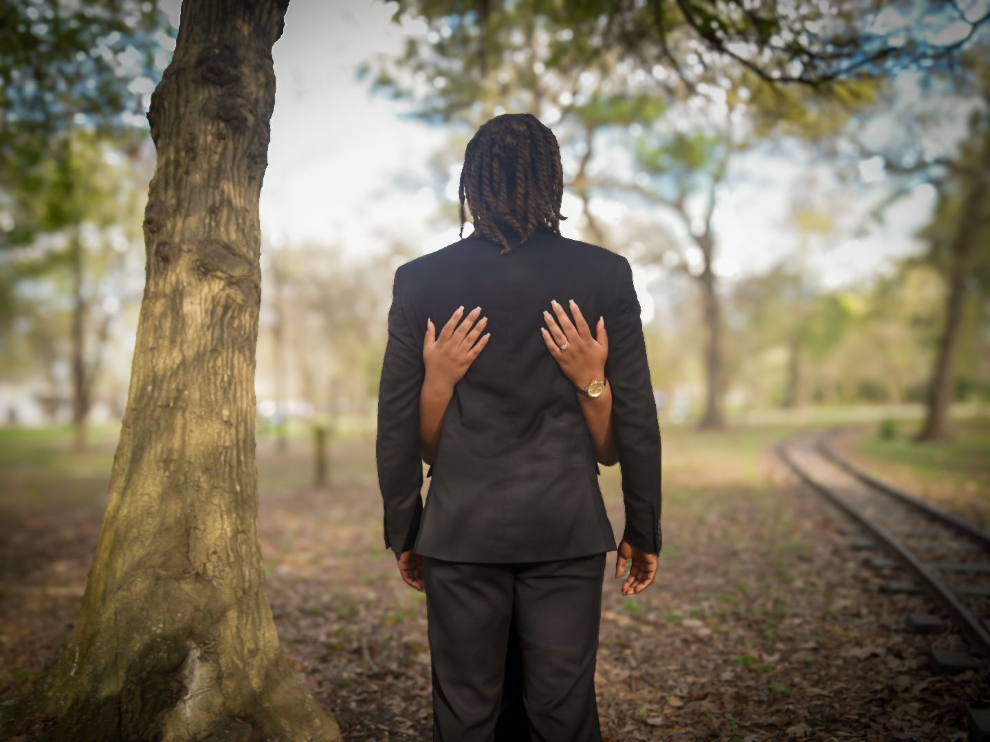 Man in black suit standing near tree by railroad tracks, with hands placed on his back.