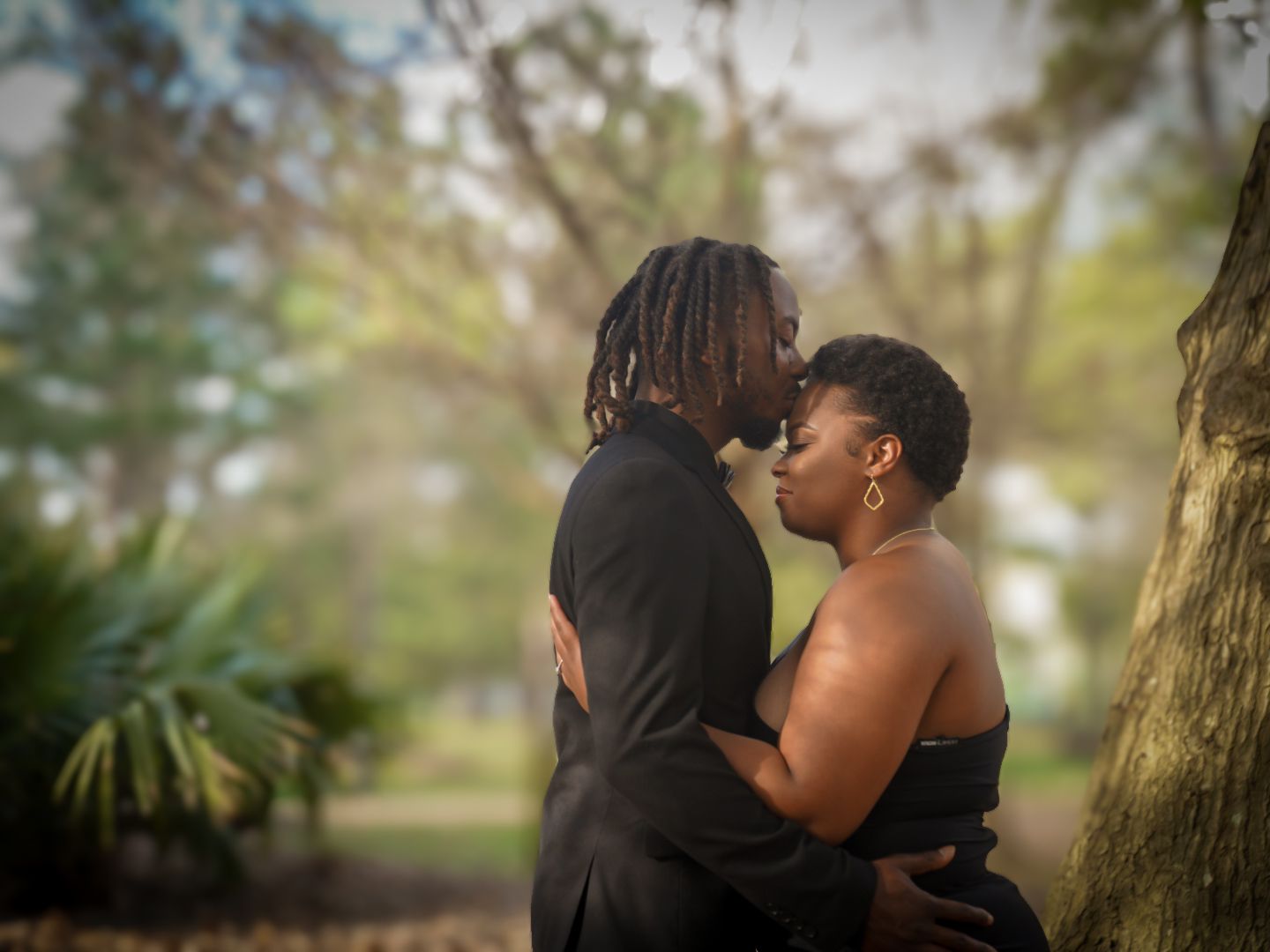 A couple shares a romantic almost-kiss embrace outdoors among trees in a lush park setting.