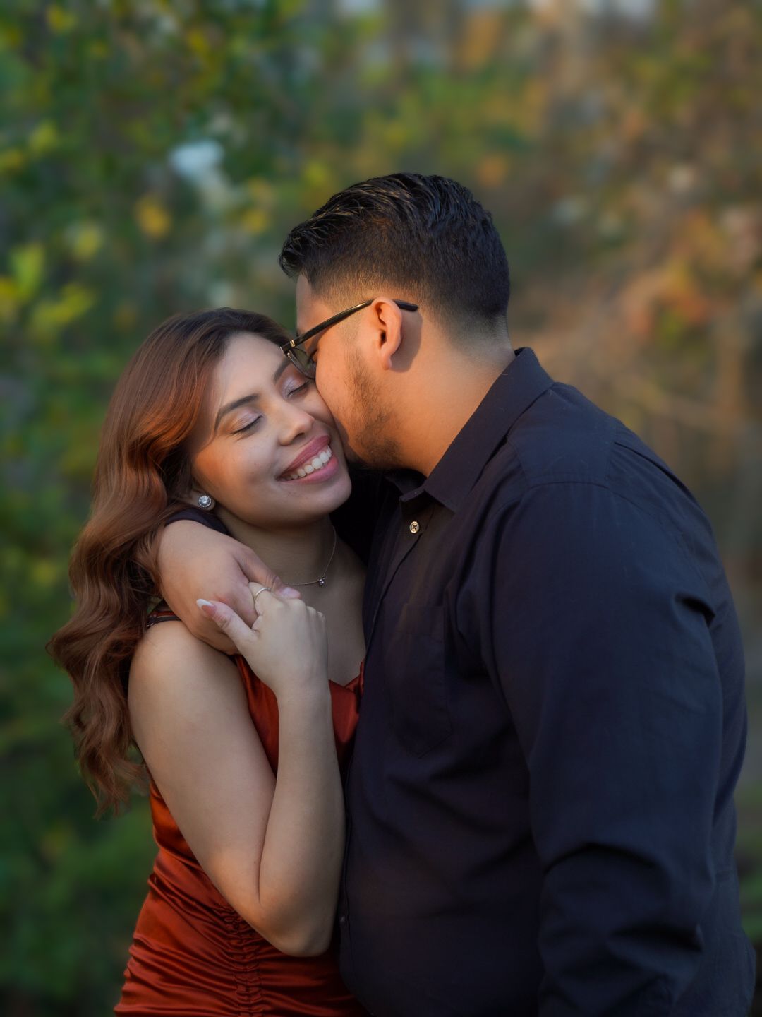 A smiling woman in a red dress embraces a man in a dark shirt outdoors with green foliage background.