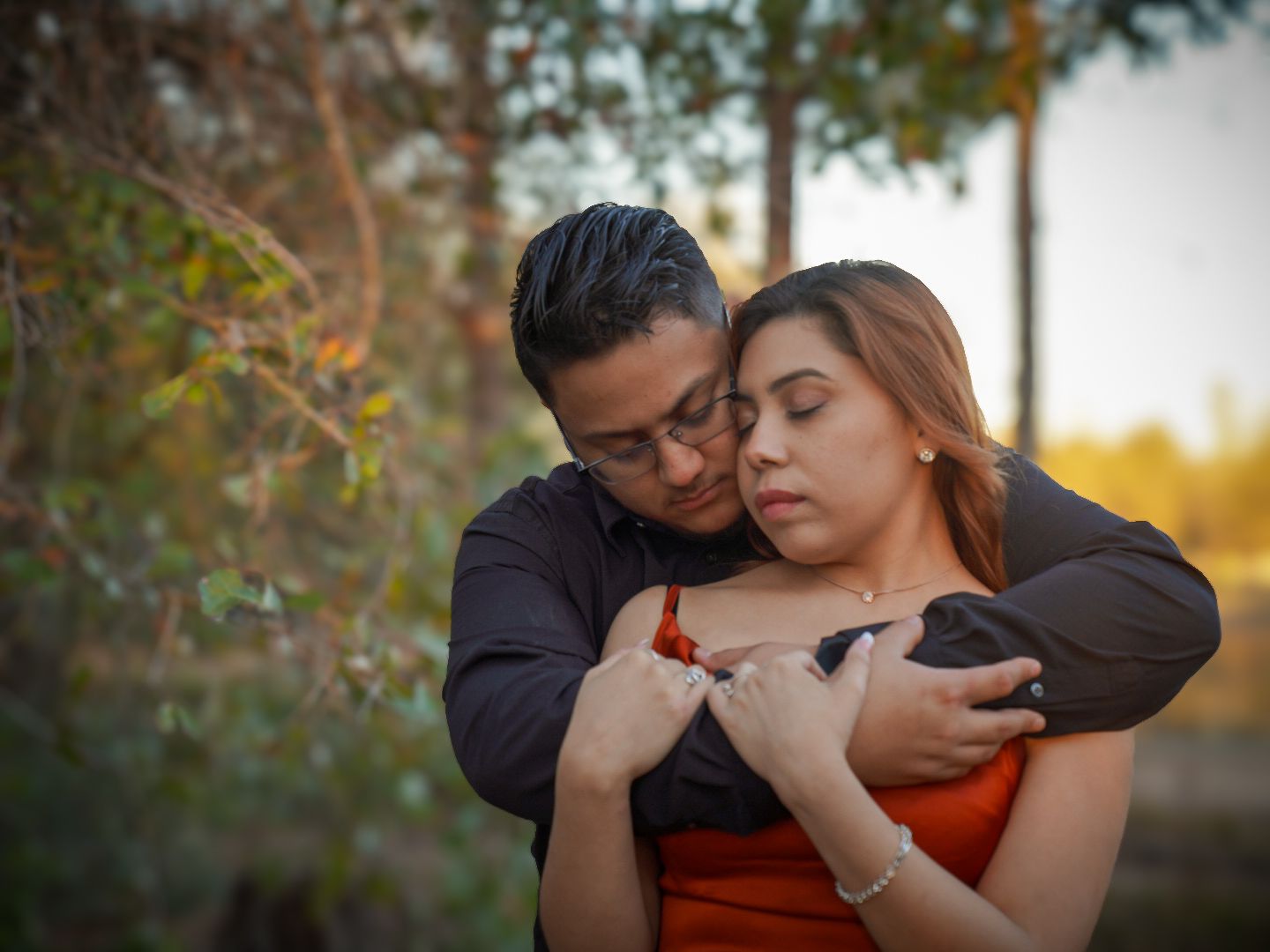 A couple embraces tenderly outdoors, man in dark jacket hugging woman in red dress among autumn trees.