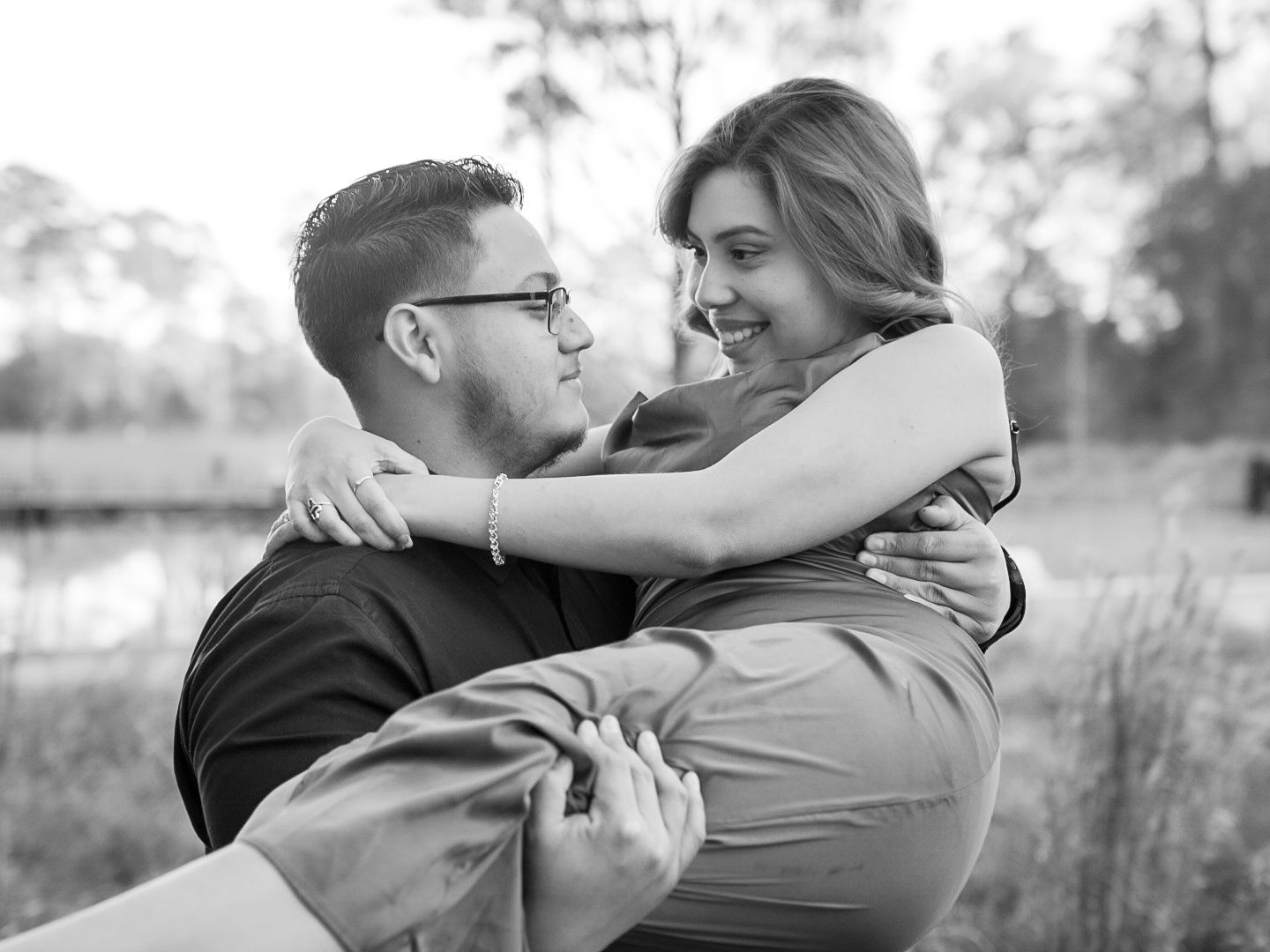 Black and white photo of a couple embracing outdoors, man lifting woman as she smiles joyfully near a pond.
