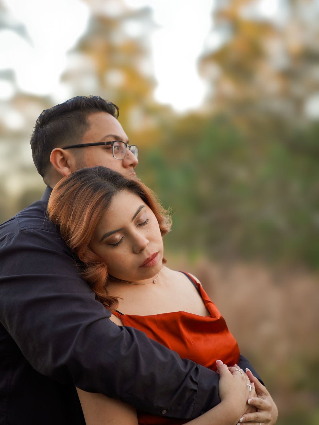 A couple shares an intimate embrace outdoors, man in dark jacket holding woman in red dress with fall foliage behind.