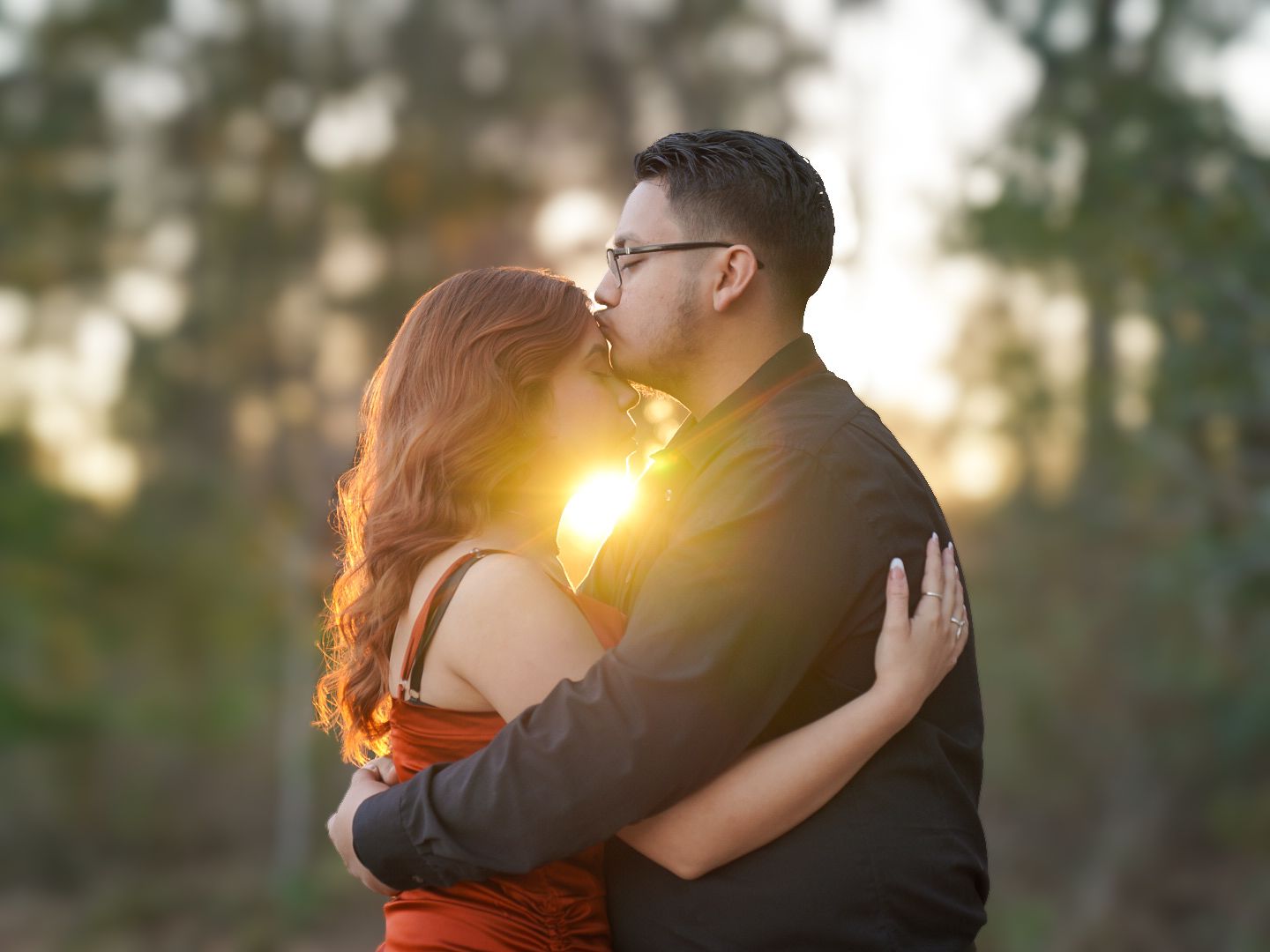 Couple sharing a romantic kiss at golden hour sunset in a lush outdoor setting, woman in red dress.